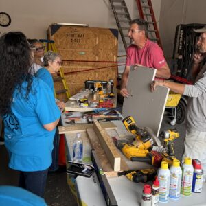 A group of people discuss tools and materials on a workbench in a workshop setting.