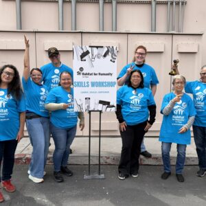 Eight people in blue Habitat for Humanity shirts smile and pose by a Skills Workshop sign outdoors.