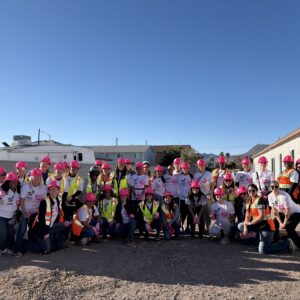 A group of people in pink hard hats and safety vests pose together at a construction site under a clear sky.
