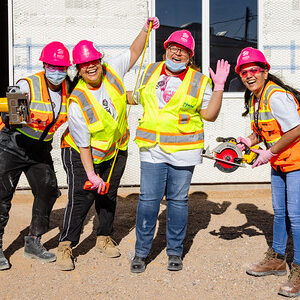 Four women in safety gear and pink hard hats pose happily with construction tools at a work site.