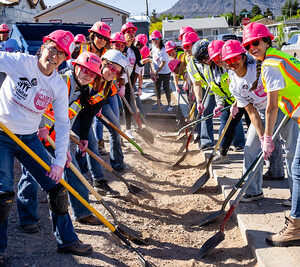 A group of people in hard hats and safety vests smile while shoveling dirt at a construction site.