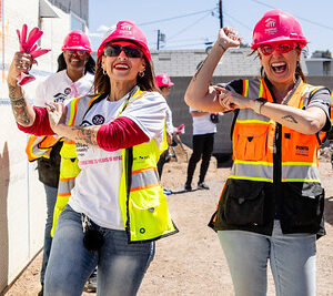 Two women in safety vests and hard hats smile and dance at a construction site with others in the background.