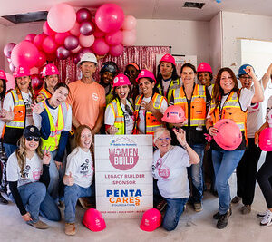 A group of women in construction gear pose around a Women Build event sign with pink balloons in the background.