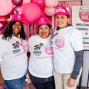 Three women in Habitat for Humanity shirts and pink hats pose smiling at a Women Build event with balloon decor.