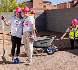 Two women in hard hats smiling with a shovel, while another woman digs near a wheelbarrow at a construction site.