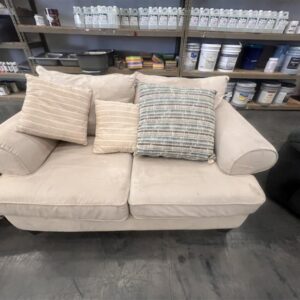 Beige loveseat with three striped pillows, displayed indoors near shelves with paint cans and supplies.