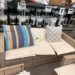 Wicker outdoor sofa with cream cushions and three decorative pillows, in a hardware store aisle.