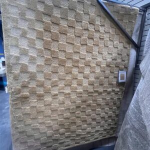 A beige rug with a raised checkerboard pattern is displayed upright in a store setting.