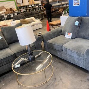 Two gray loveseats, a round glass coffee table, and a lamp displayed in a furniture store.