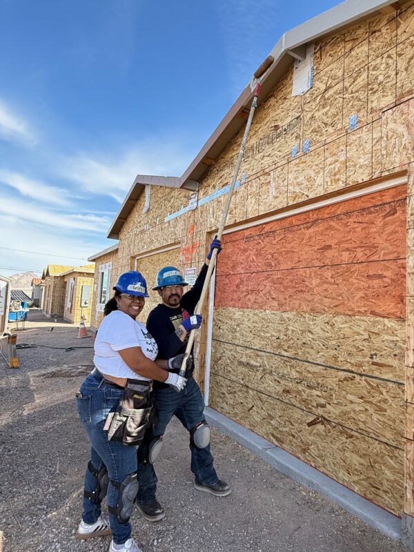 Two people in hard hats smile while painting the exterior of a house under construction.