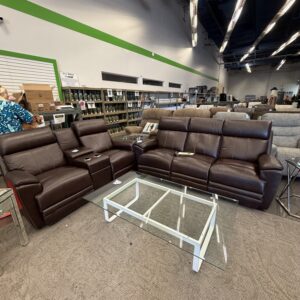 Brown leather sofas and a glass coffee table are displayed in a furniture store with shelves in the background.