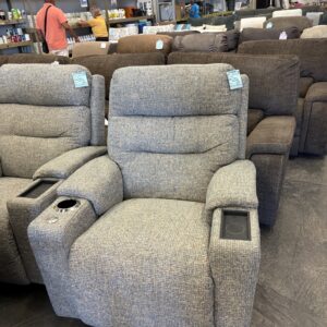 Gray recliner chair with cup holders on display in a furniture store, other chairs and shoppers in the background.