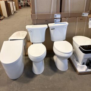 Four different toilets are displayed side by side on a warehouse floor with boxes and shelves in the background.