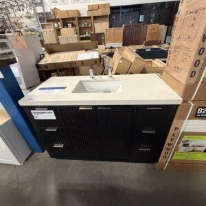 A black vanity with a white sink top is displayed in a store among boxes and other cabinets.