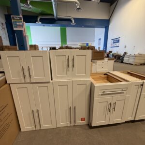White kitchen cabinets on display in a warehouse, with some drawers open and sale signs visible in the background.