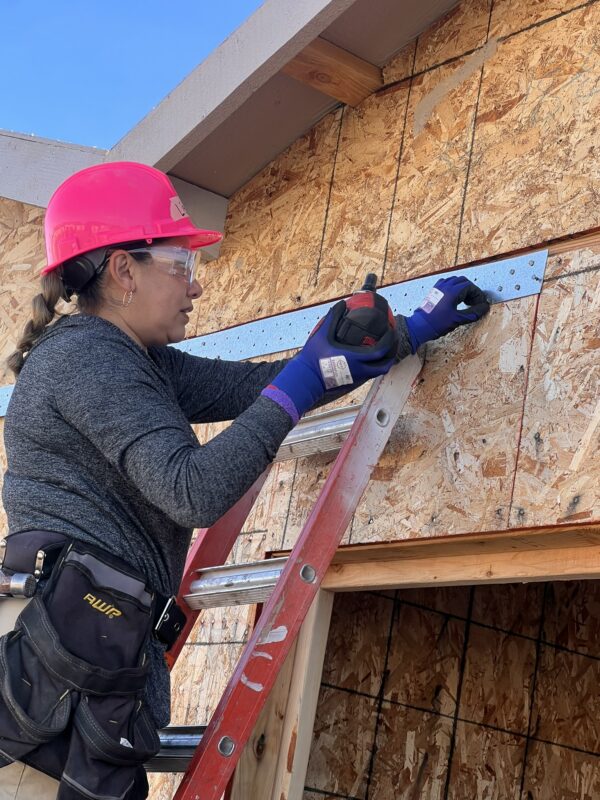 Woman in safety gear on ladder using a drill to secure a metal plate to a wooden building under construction.