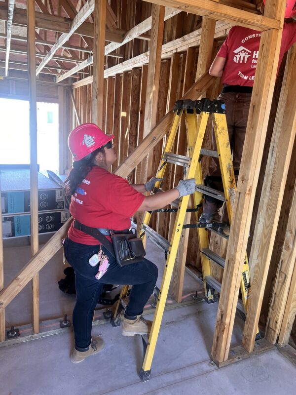 Two people in red shirts and hard hats work on a wooden house frame; one climbs a yellow ladder.