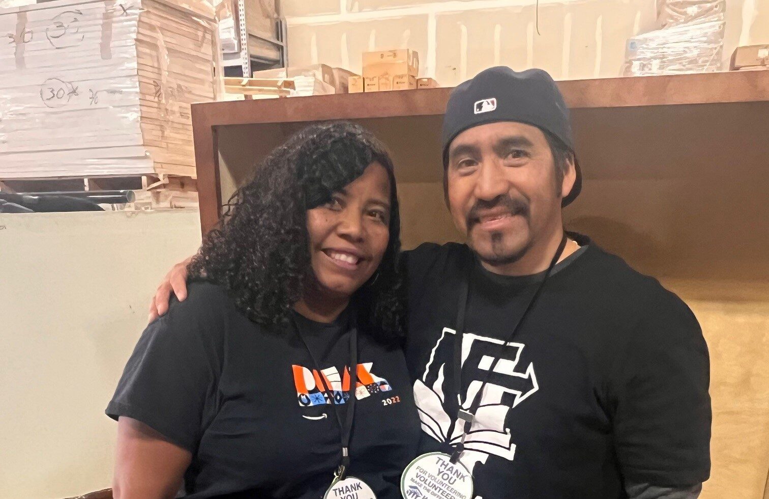 Two people smiling and standing together in a warehouse, both wearing black shirts and event lanyards.