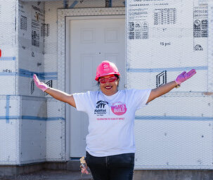 Woman in pink hard hat and gloves smiles with arms outstretched in front of a house under construction.