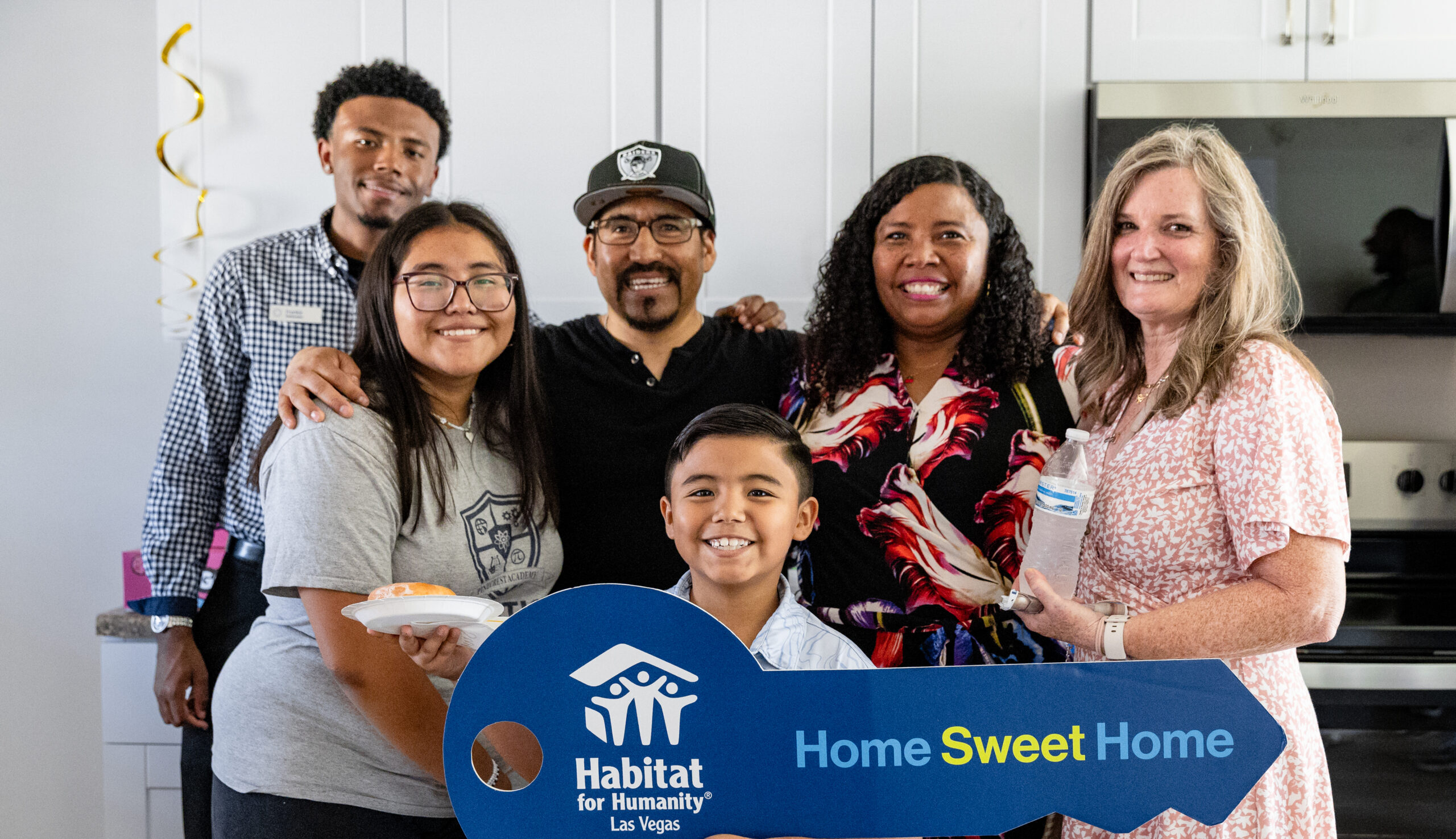 Six people smile together in a kitchen, holding a large Home Sweet Home Habitat for Humanity key.