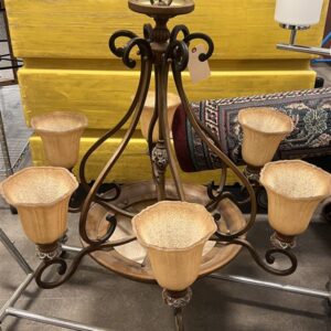 A bronze chandelier with five frosted glass shades sits on a metal shelf near a rolled-up rug and a lamp.