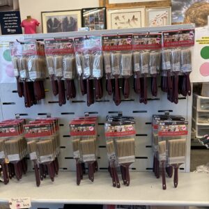 Paintbrushes in plastic packaging are displayed on hooks on a store shelf with price tags visible below.