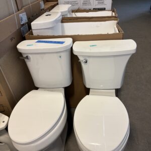 Two white toilets displayed side by side in a store, with boxes and other toilets in the background.