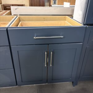 Dark blue kitchen cabinets with silver handles, missing the countertop, displayed in a warehouse setting.