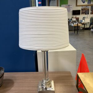A modern table lamp with a white ribbed shade and silver base sits on a wooden table in a store.