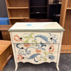 A white dresser with three drawers, decorated with colorful floral designs, sits among wooden shelves.
