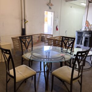 Glass round dining table with four chairs in a warehouse room, bench and exit door in the background.