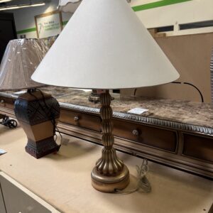 Gold table lamp with a white shade on a wooden surface, next to another lamp and a dresser in a store.