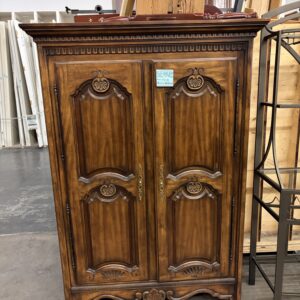 Ornate wooden armoire with decorative carvings, double doors, and brass handles in a warehouse setting.