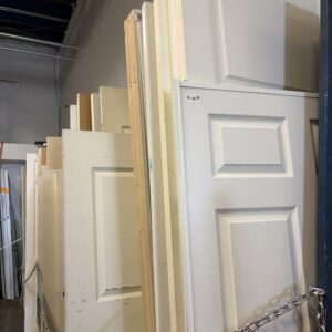 A row of standing white doors is displayed behind a metal chain in a warehouse setting.