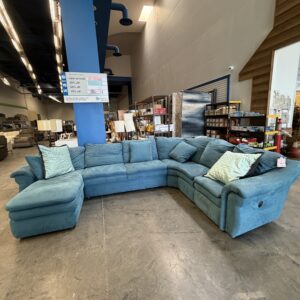 Large teal sectional sofa with two pillows in a furniture store, displayed on a polished concrete floor.
