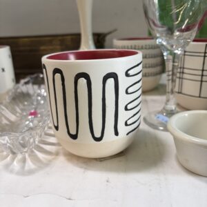 A white ceramic cup with black wavy lines sits among assorted glassware and dishes on a shelf.