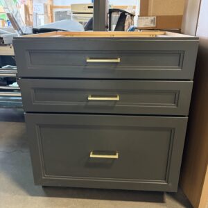 A dark gray cabinet with three drawers and gold handles sits on a concrete floor in a store.