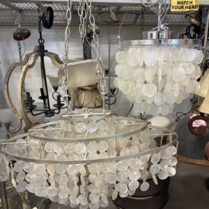 Two hanging chandeliers with white capiz shells are displayed below a yellow caution sign in a store.