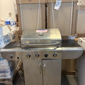 A stainless steel gas grill with four burners on display in a store, surrounded by boxes and cabinets.