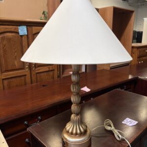 Table lamp with a white shade and a gold decorative base on a wooden desk in a furniture store.
