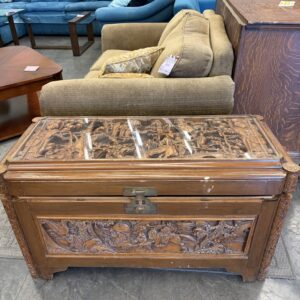 Carved wooden chest with brass lock sits in front of a tan loveseat in a secondhand furniture store.
