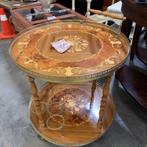 Ornate wooden serving cart with floral inlay, two shelves, and a note on top, in a thrift store setting.