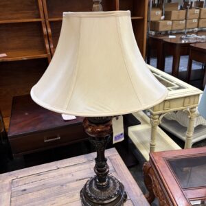 A vintage table lamp with a cream lampshade sits on a wooden table in a thrift store.