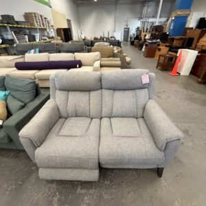 Gray two-seat reclining sofa displayed in a furniture store with other couches and shelves in the background.