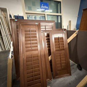 Five wooden window shutters are stacked in front of a large window inside a workshop.