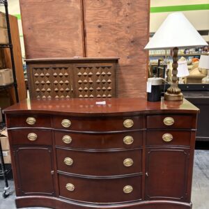 A dark wood dresser with gold handles, a brass lamp, and two wooden cabinets behind it in a store.