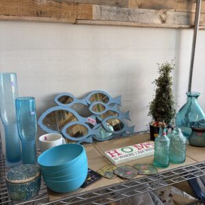 Blue glassware, bowls, fish-shaped mirrors, and home decor items arranged on a metal shelf against a wood wall.