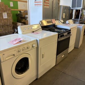 A row of used laundry and kitchen appliances for sale in a store, including washers, dryers, and an oven.