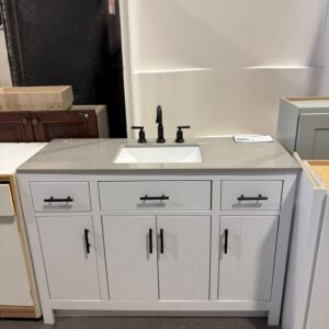 White bathroom vanity with a gray countertop, black faucet, and three drawers, displayed in a showroom.