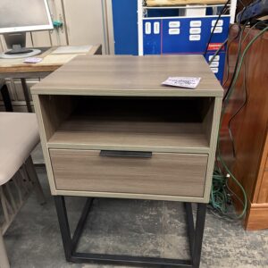 Modern wood nightstand with open shelf and drawer, sitting on a black metal base in a store setting.